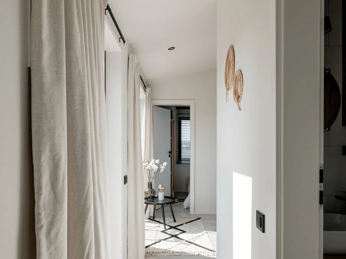 Een helder en minimalistisch ingerichte gang in een klein huis met natuurlijke linnen gordijnen, een kleine bijzettafel met decoratieve elementen en kunstzinnig geplaatste wanddecoraties.