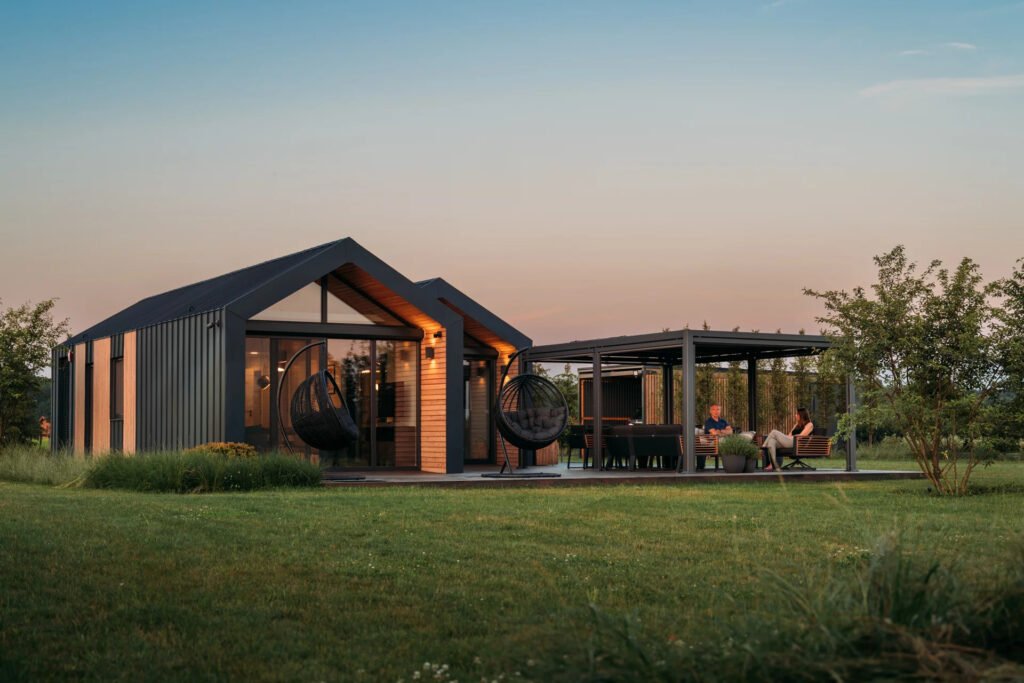 Een moderne modulaire woning tijdens de schemering, met een gezellig verlicht interieur en een overdekte buitenruimte waar twee mensen ontspannen praten.