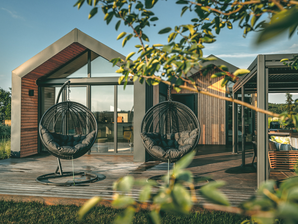 Een sfeervol buitenterras van een moderne mantelzorgwoning met grote hangende rotan stoelen, houten bekleding, een glazen gevel en een pergola, omringd door weelderig groen.