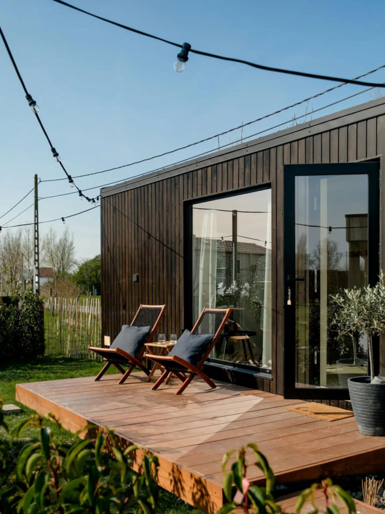 Buitenkant van een modern houten huis met een groot terras, voorzien van loungestoelen en omgeven door een tuin en sfeerverlichting