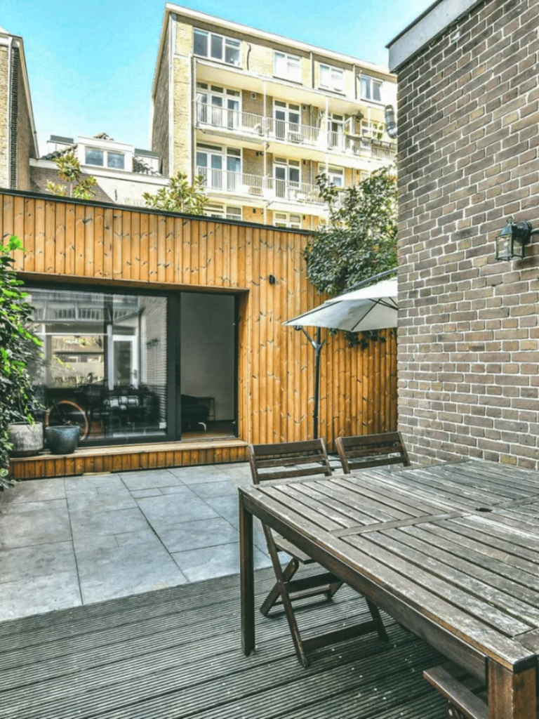 Besloten achtertuin met een groot houten terras en een eettafel, omsloten door bakstenen muren en een huis met houten gevel