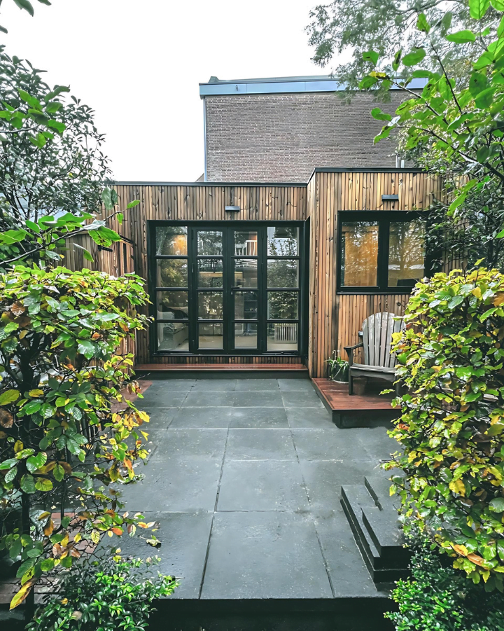 Een modern tiny house met grote glazen deuren en ramen, omgeven door groen, met een ruime stenen patio en een houten stoel