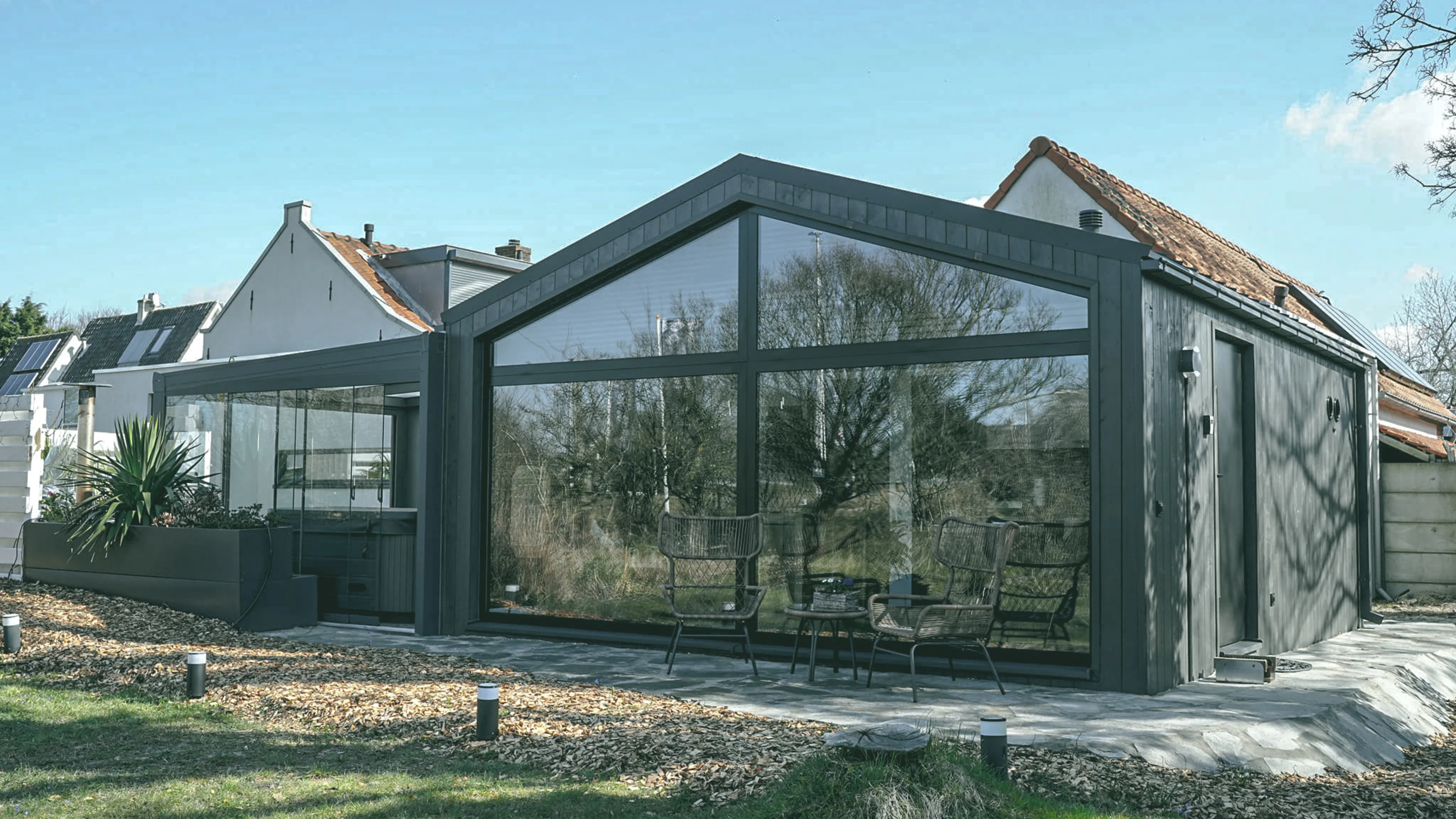 Een moderne, custom ontworpen tiny home met grote glazen panelen aan de zijkant van een klassieke woning met een schuin dak. De serre is verbonden met een terras waarop donkergrijze tuinstoelen staan. De setting toont een samensmelting van moderne architectuur en traditionele woningbouw, omgeven door een rustige tuin.