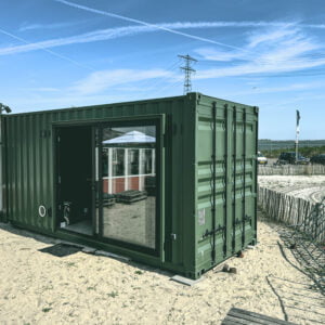 Op de afbeelding zie je een containerwoning, gemaakt van een groene zeecontainer. De container is geplaatst op een zanderig terrein, waarschijnlijk in een buitenomgeving. De woning heeft een grote glazen schuifdeur die toegang biedt tot het interieur. Aan de linkerkant van de container zijn ventilatieopeningen zichtbaar. Op de achtergrond zie je een omheining, enkele geparkeerde auto’s en een vlag. De lucht is helder blauw met enkele vliegtuigstrepen. Het lijkt een zonnige dag te zijn.