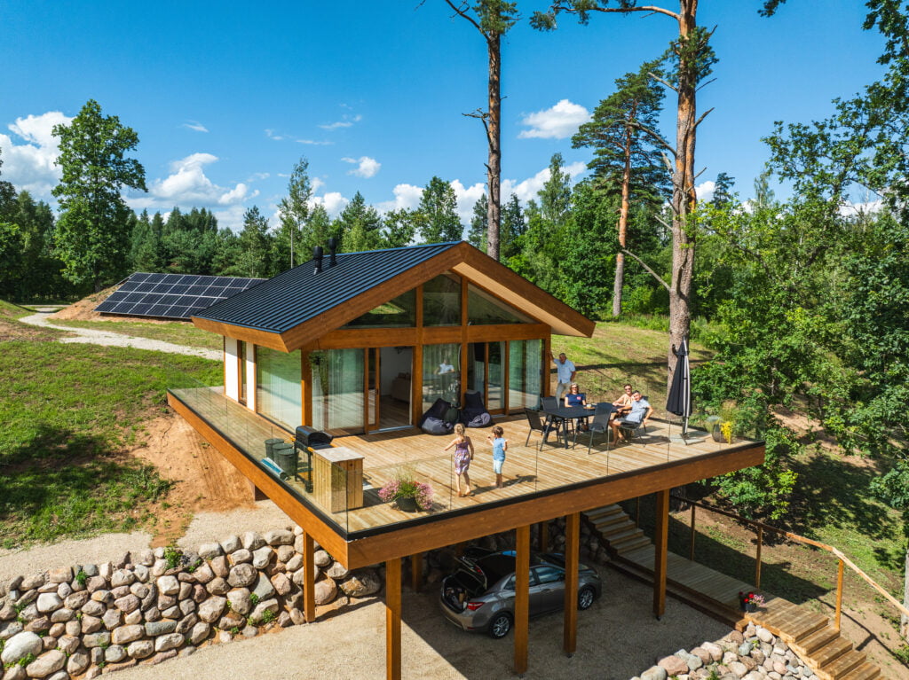 Een CLT houten huis gefotografeerd vanuit de lucht op een zonnige dag. Het huis heeft grote glazen ramen en een ruim terras, waar een gezin samen zit aan een eettafel en twee kinderen spelen op het dek. Onder het verhoogde huis is een carport met een geparkeerde auto. Op de achtergrond zijn zonnepanelen te zien op een zonnige open plek in een bosrijke omgeving. Het huis en de omgeving stralen duurzaamheid en rust uit.