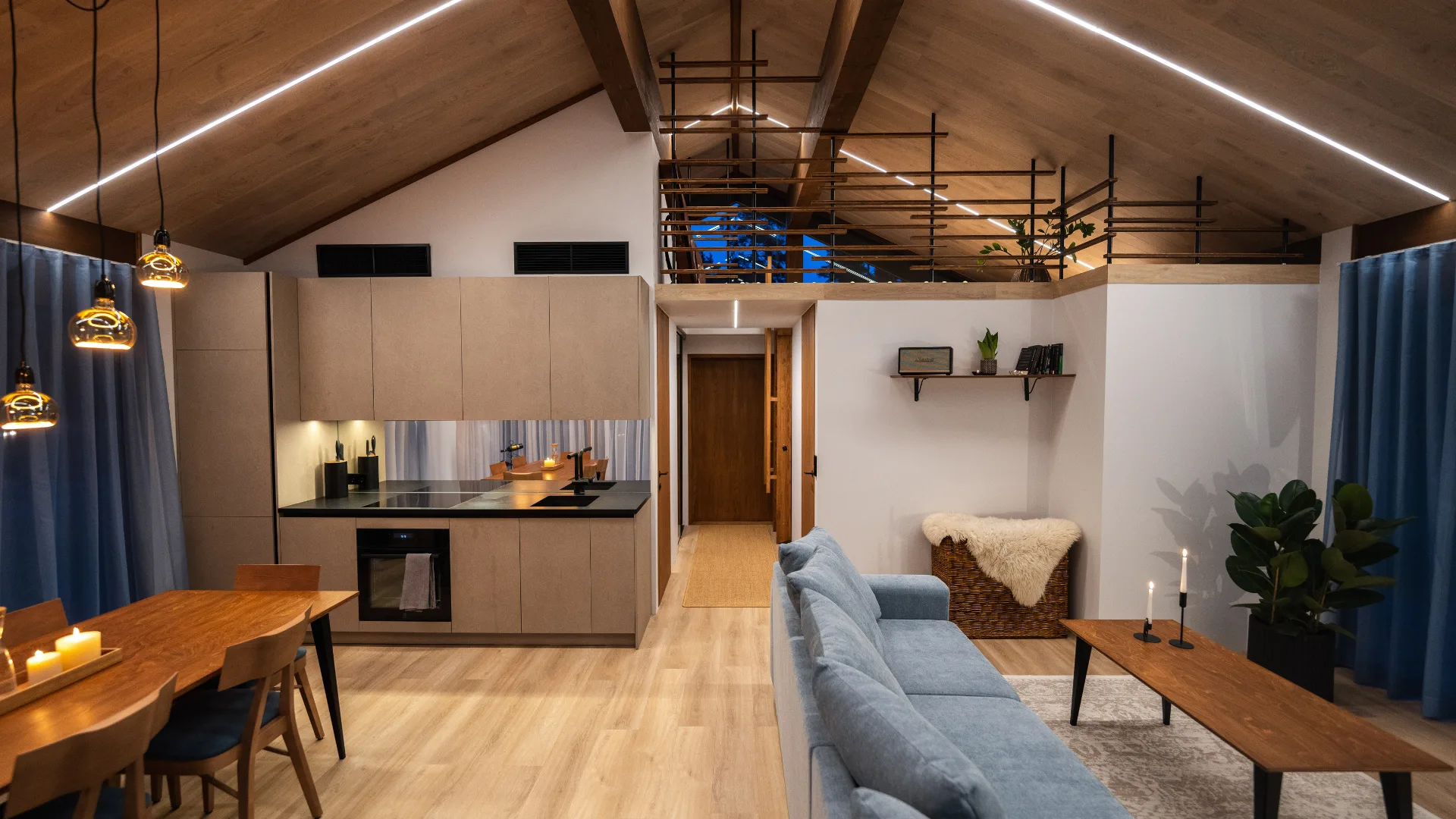 Modern interieur van een houten huis met een open keuken en zitruimte. De ruimte is ingericht met een blauwe bank, houten meubels en sfeerverlichting onder een hoog, houten plafond.