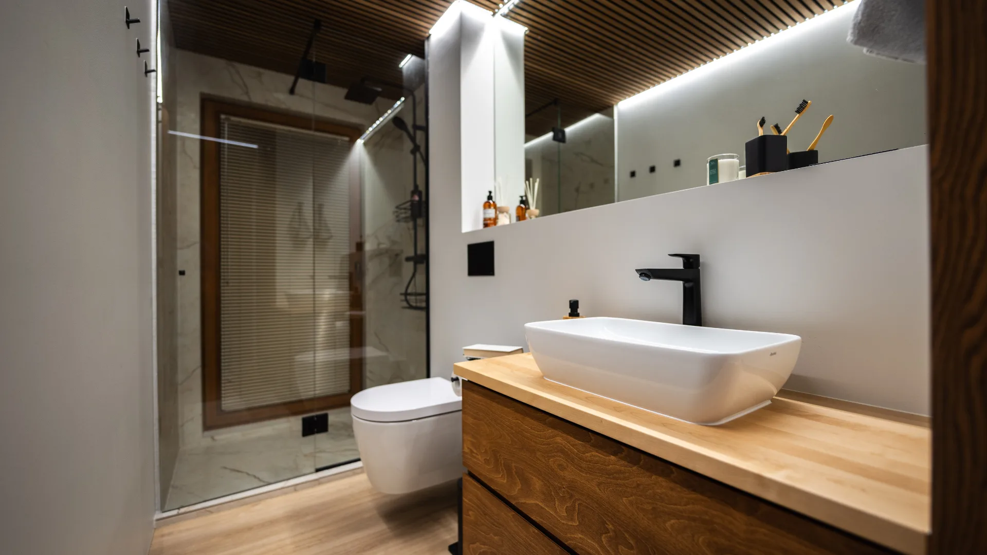 Moderne badkamer in een houten huis, voorzien van een glazen douche, een zwevend toilet en een wastafel met houten kastje. Zachte verlichting zorgt voor een warme sfeer in de ruimte.