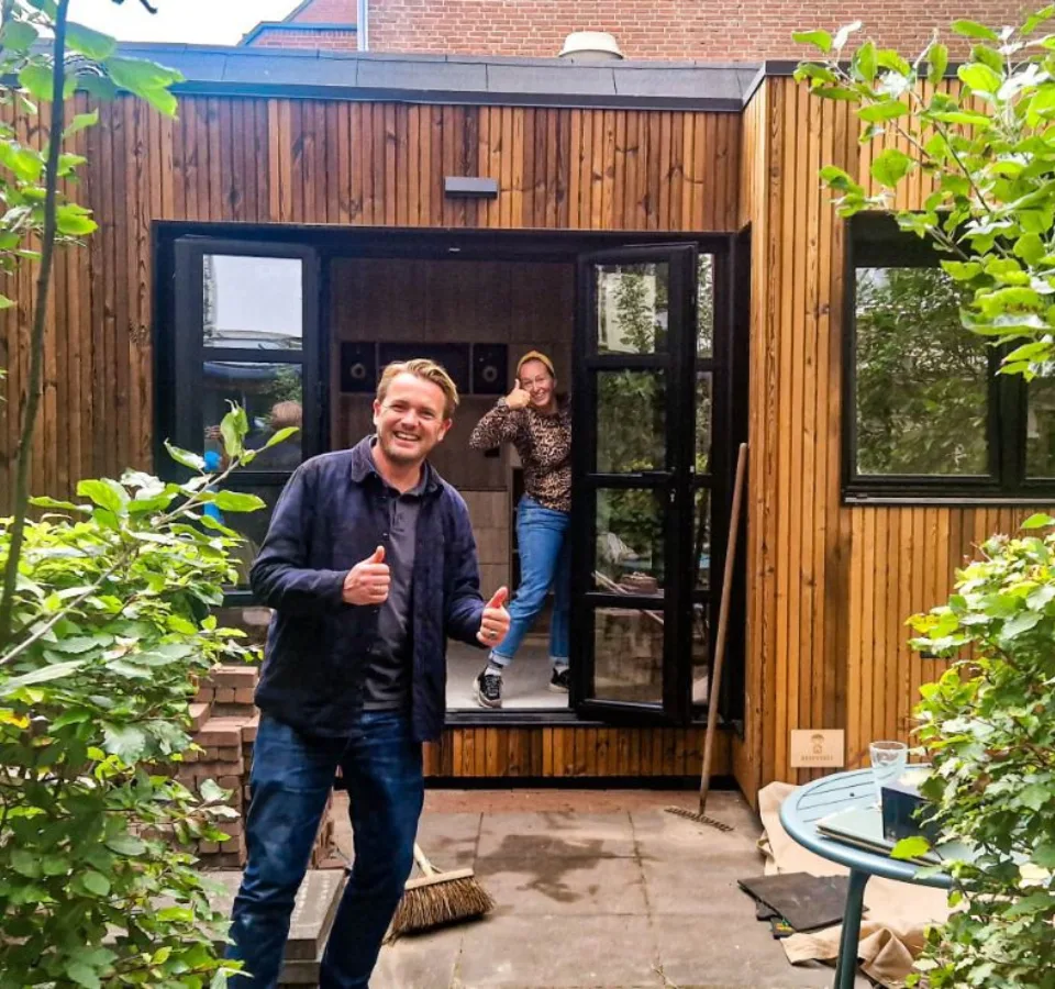 Twee tevreden klanten poseren enthousiast voor hun op maat gemaakte tiny house met houten gevel. De man staat buiten en steekt zijn duimen omhoog, terwijl de vrouw vanuit de deuropening glimlacht en een duim omhoog steekt. Het tiny house heeft een moderne uitstraling met grote ramen en een strak design, omgeven door groen en een betegelde tuin.