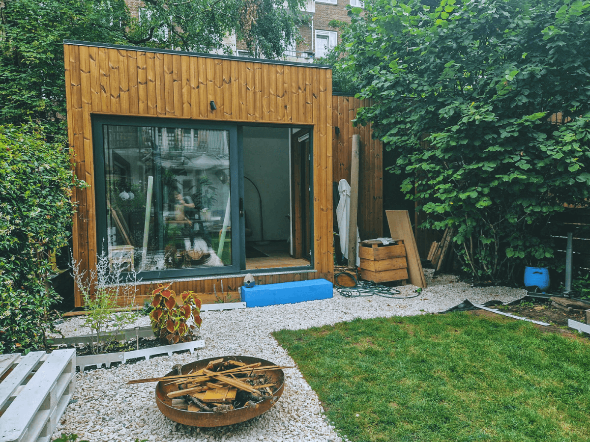 Een modern houten tiny house met grote glazen schuifdeuren staat in een groene achtertuin, omringd door bomen en planten. De tuin heeft een vuurplaats, grindpaden en enkele bouwmaterialen, wat wijst op een recente plaatsing of afwerking van de woning.