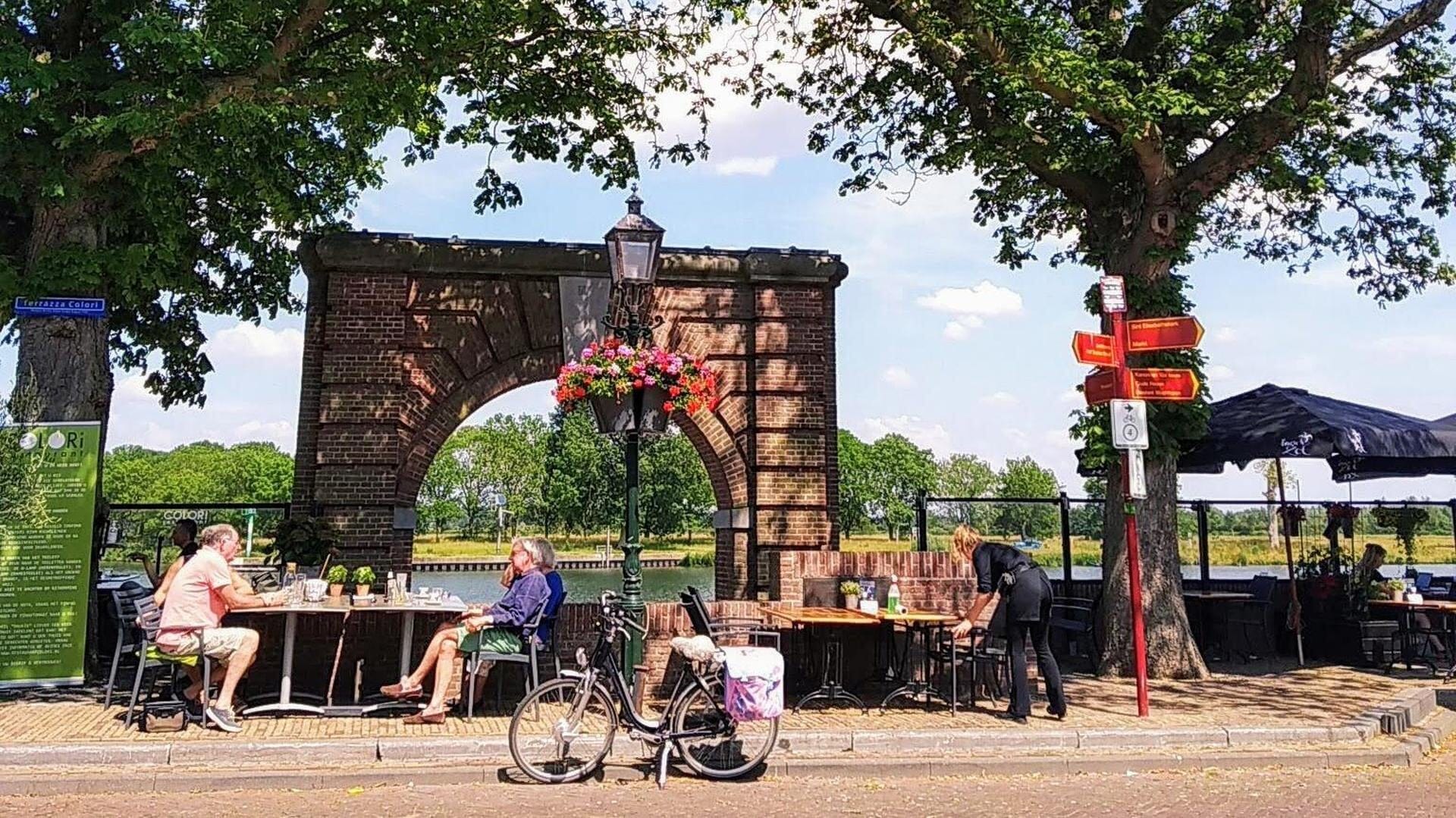 Gezellig terras met mensen die genieten van een zonnige dag in Land van Cuijk, met een historische stadspoort en groene omgeving op de achtergrond.