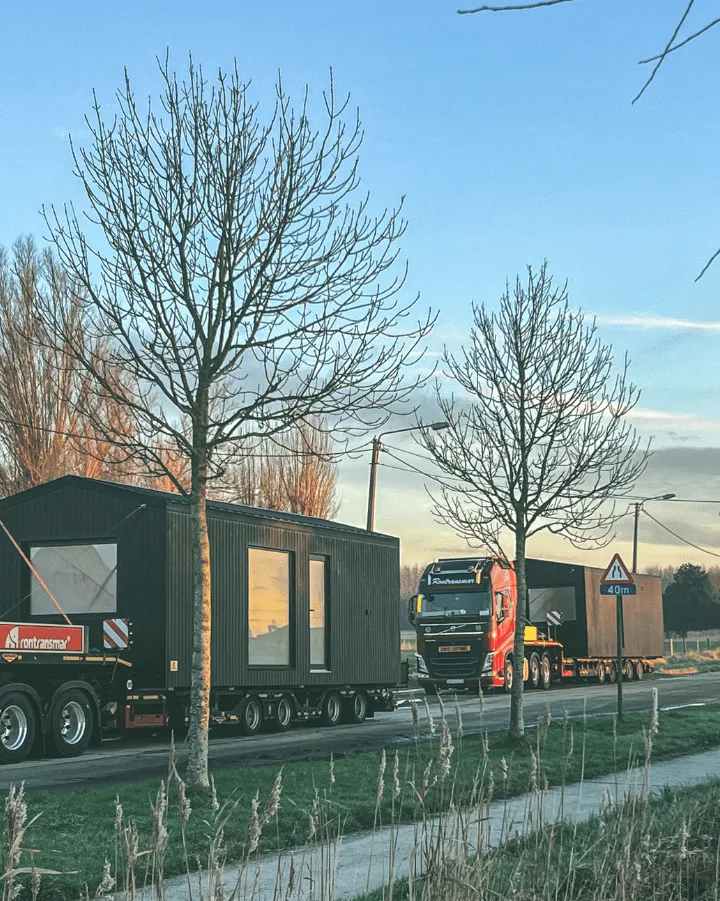 Tiny House op speciaal transport onderweg naar nieuwe locatie, begeleid door vrachtwagen bij zonsondergang