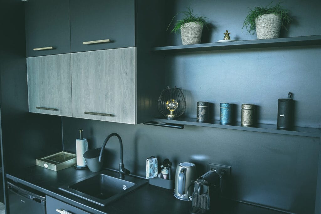 Moderne zwarte keuken met licht houten bovenkasten, zwarte spoelbak, matzwarte kraan en ingebouwde koffie- en waterkoker, decoratief verlicht met een designlamp op de plank.