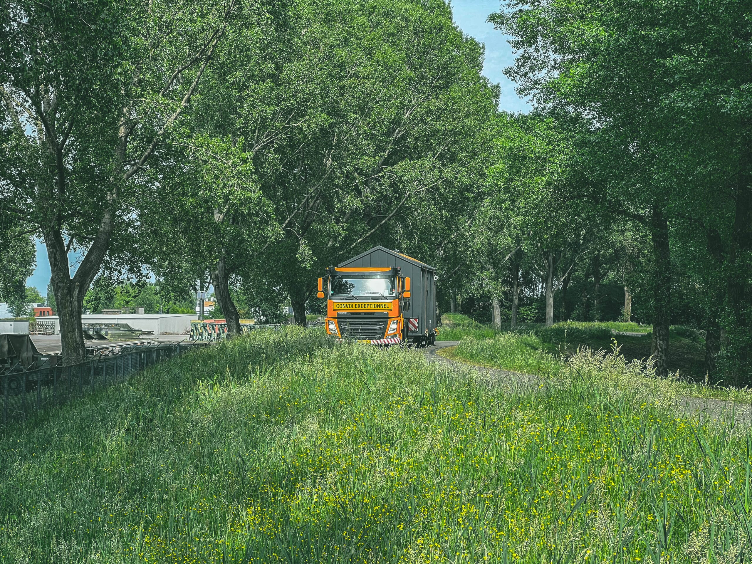 Transport van een Tiny House via Convoi Exceptionnel vrachtwagen door een groen landschap met hoge bomen.