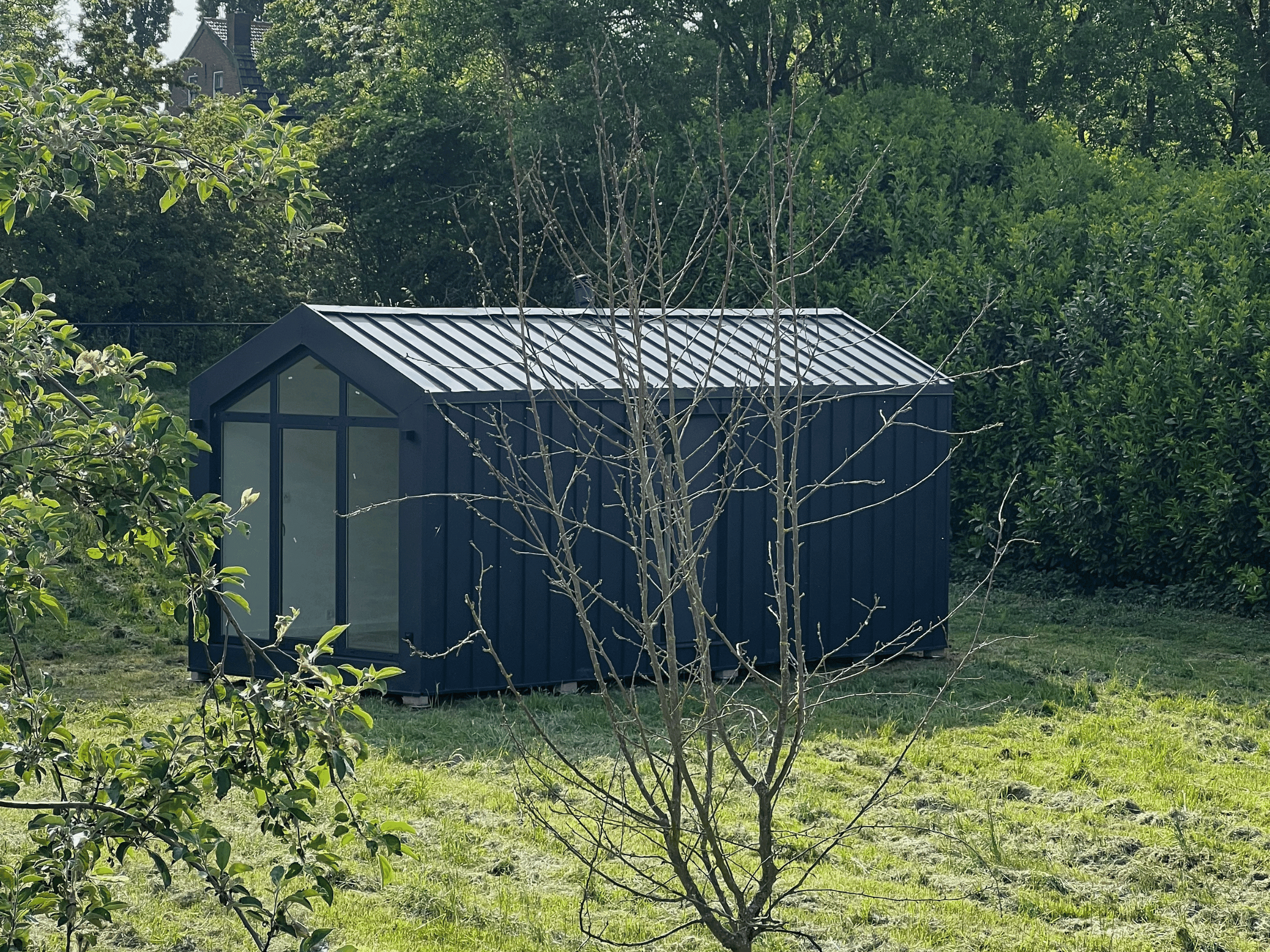 Tiny House geplaatst in een groene weide, met strak zwart exterieur en glazen gevel, omringd door bomen en een pas gemaaid grasveld.