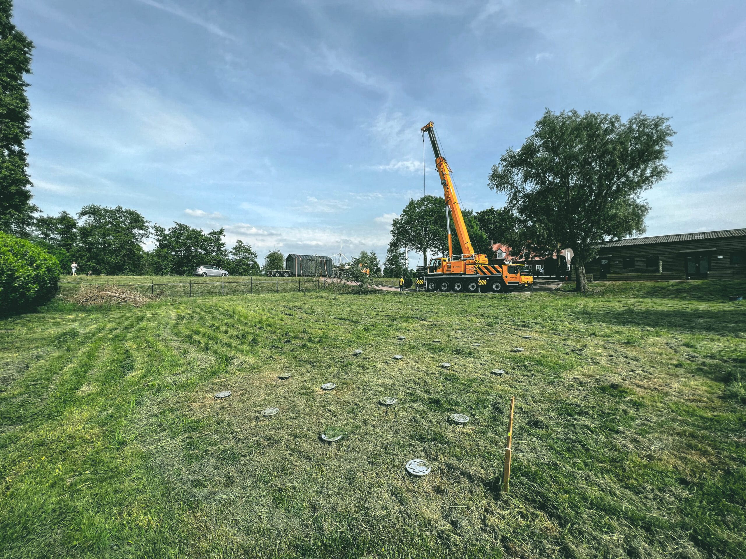 Grote hijskraan plaatst een Tiny House op schroeffundering in een open veld met zicht op het erf en omliggende gebouwen.