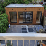 Compacte houten familiewoning met schuifdeuren in een achtertuin, omringd door groen en een betegeld terras met tuinmeubels. Ideaal als mantelzorgwoning of jongerenwoning