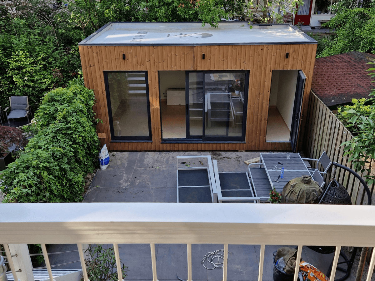 Compacte houten familiewoning met schuifdeuren in een achtertuin, omringd door groen en een betegeld terras met tuinmeubels. Ideaal als mantelzorgwoning of jongerenwoning