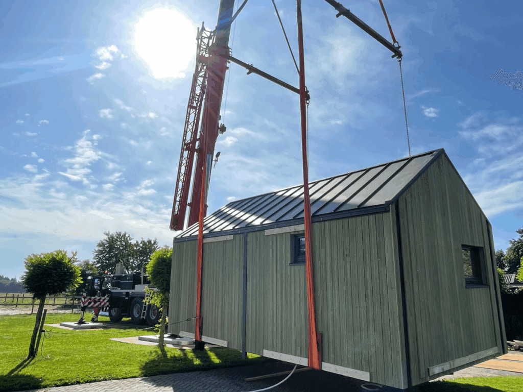 Een groen houten Tiny House wordt met een hijskraan op zijn plek gehesen op een zonnige dag. De kraan staat op het grasveld, omringd door kleine boompjes en een landelijke omgeving.
