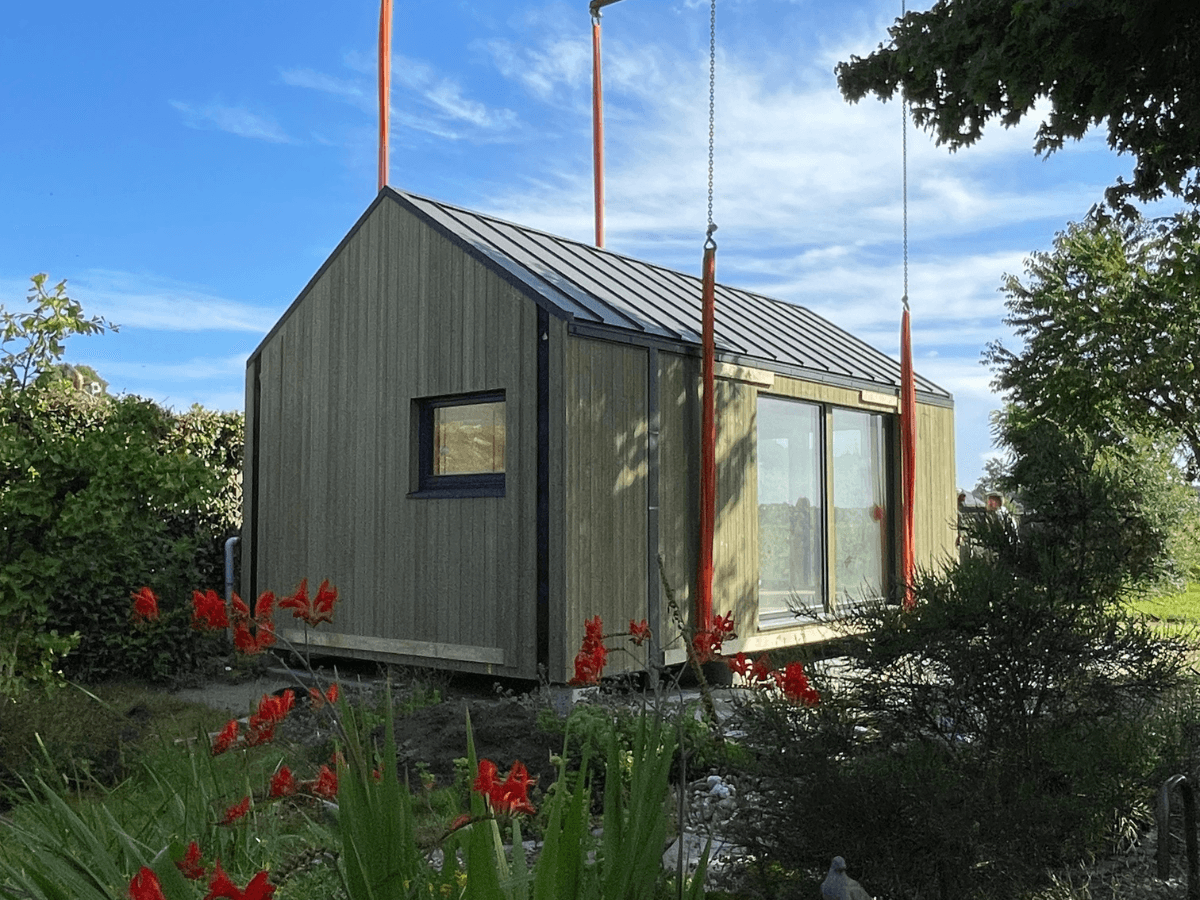 Een geplaatst groen houten Tiny House met schuifdeuren en zwart stalen dak, omringd door bloeiende planten en bomen. De hijsbanden van de kraan hangen nog om de woning
