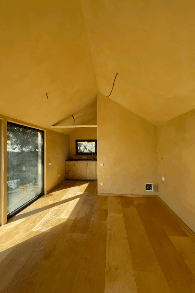 Licht en warm interieur van de tuinstudio met leemstuc wanden, houten vloer en groot raam met uitzicht op de tuin.